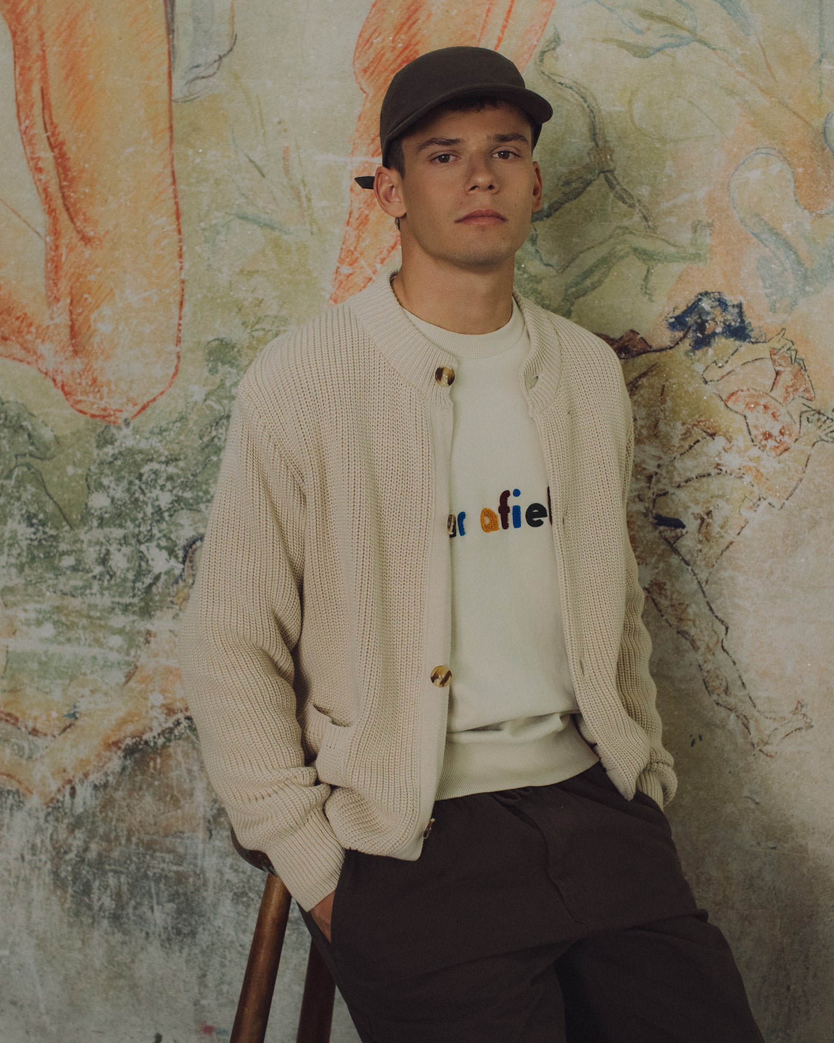 Model seated against a textured wall in a cream knit cardigan, logo sweatshirt, brown trousers and cap